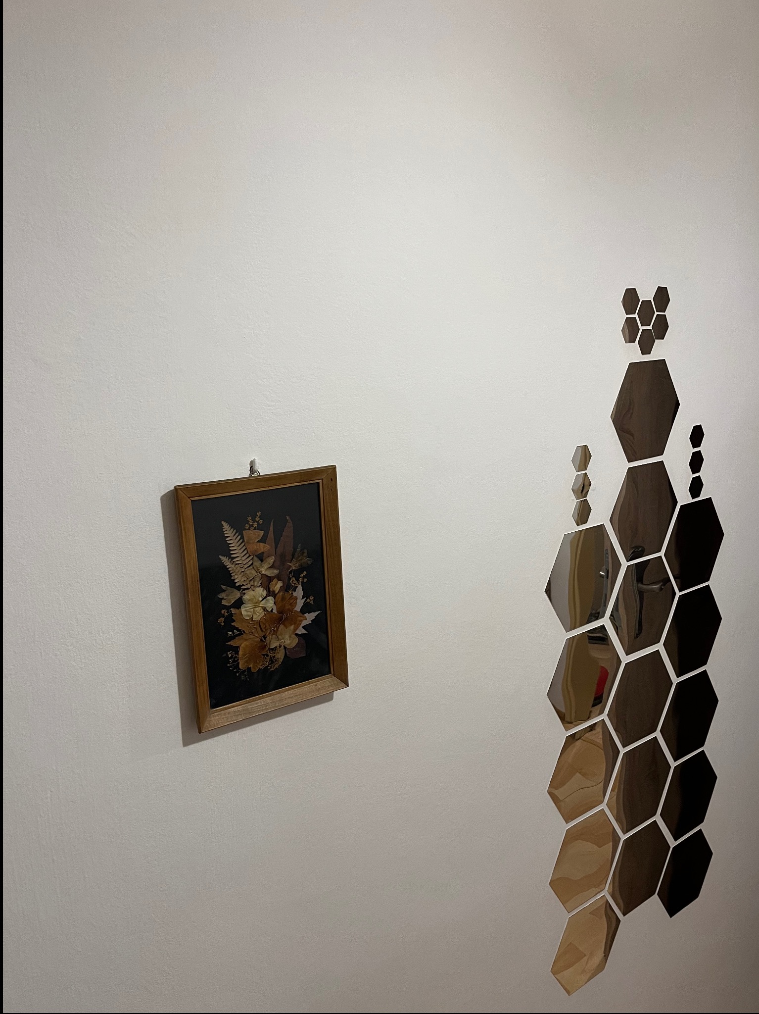 Biała ściana z dekoracją: oprawiony obraz z suszonych kwiatów i lustrzane heksagony ułożone w pionowy wzór. Minimalistyczny i osobisty akcent w aranżacji wnętrza.