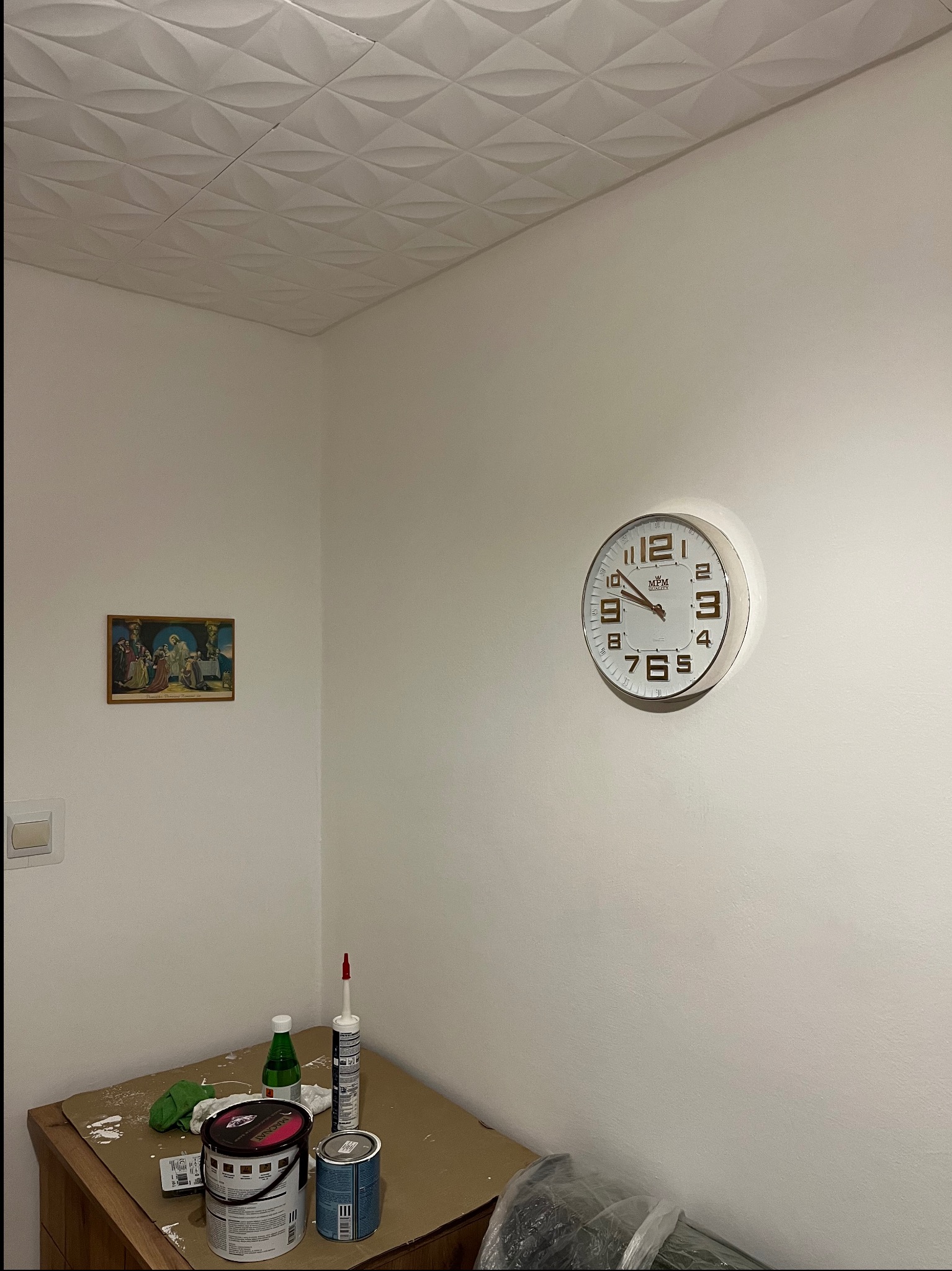 Pokój po malowaniu: białe ściany, ozdobny sufit, zegar na ścianie, obrazek i farby na komodzie. Widok na róg pokoju po odświeżeniu.