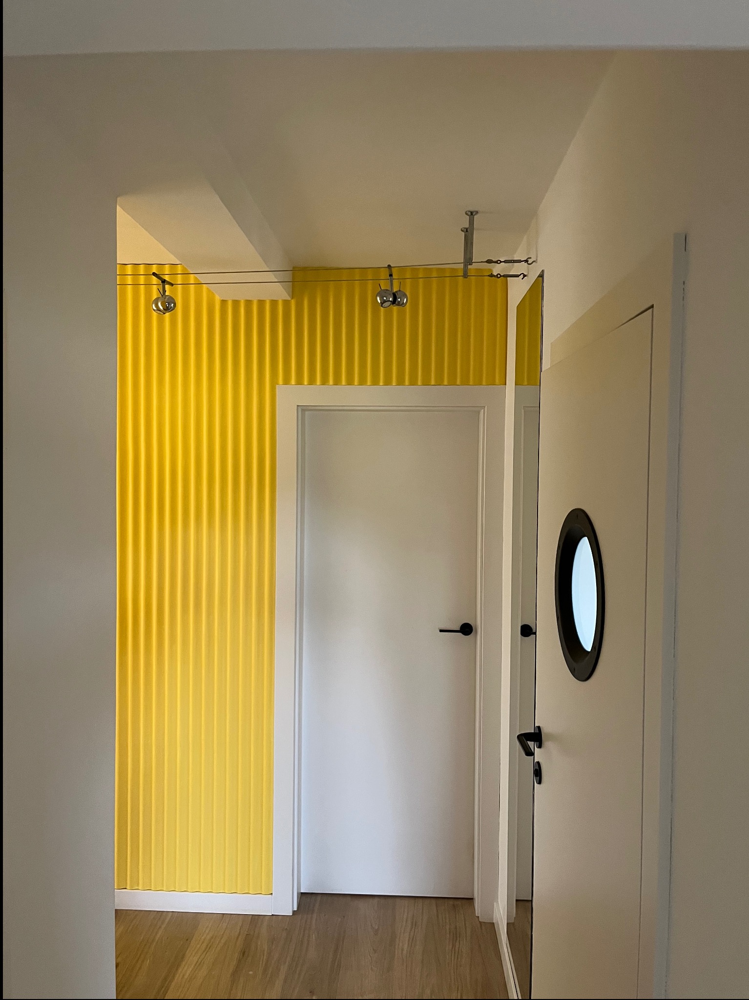 Wnętrze korytarza z żółtą, teksturowaną ścianą i białymi drzwiami z okrągłym okienkiem. Widoczne elementy oświetlenia na suficie i drewniana podłoga.