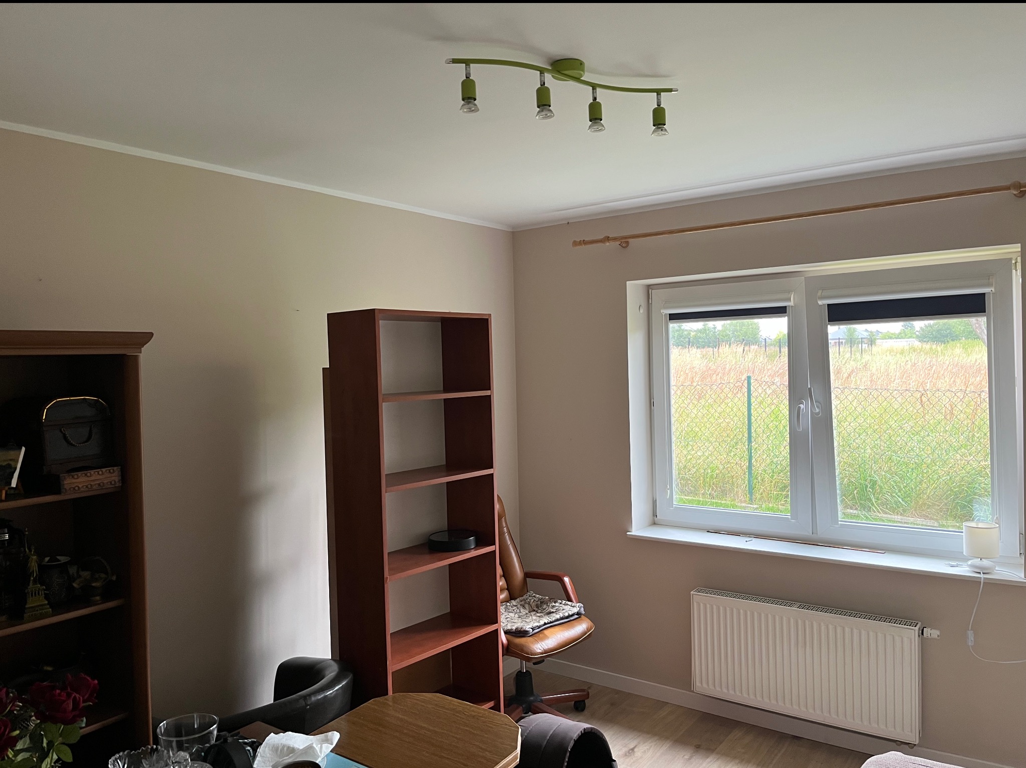 Beżowe wnętrze pokoju z brązowym regałem, biurkiem, krzesłem i białym grzejnikiem pod oknem. Ściany świeżo pomalowane na jednolity kolor.