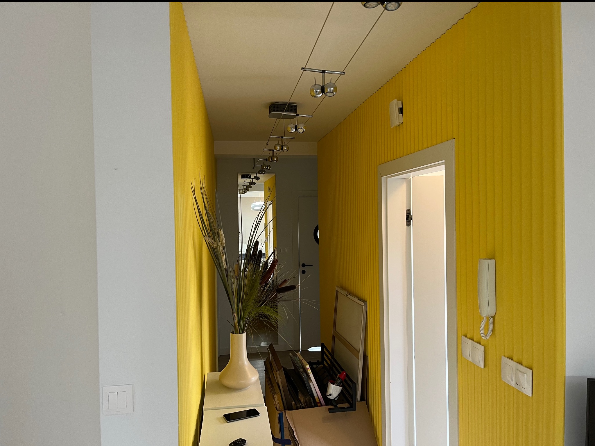 Żółty korytarz z dekoracyjnym oświetleniem, wazonem i białymi drzwiami. Ściany w pionowe pasy. Widoczne detale wykończenia wnętrza.