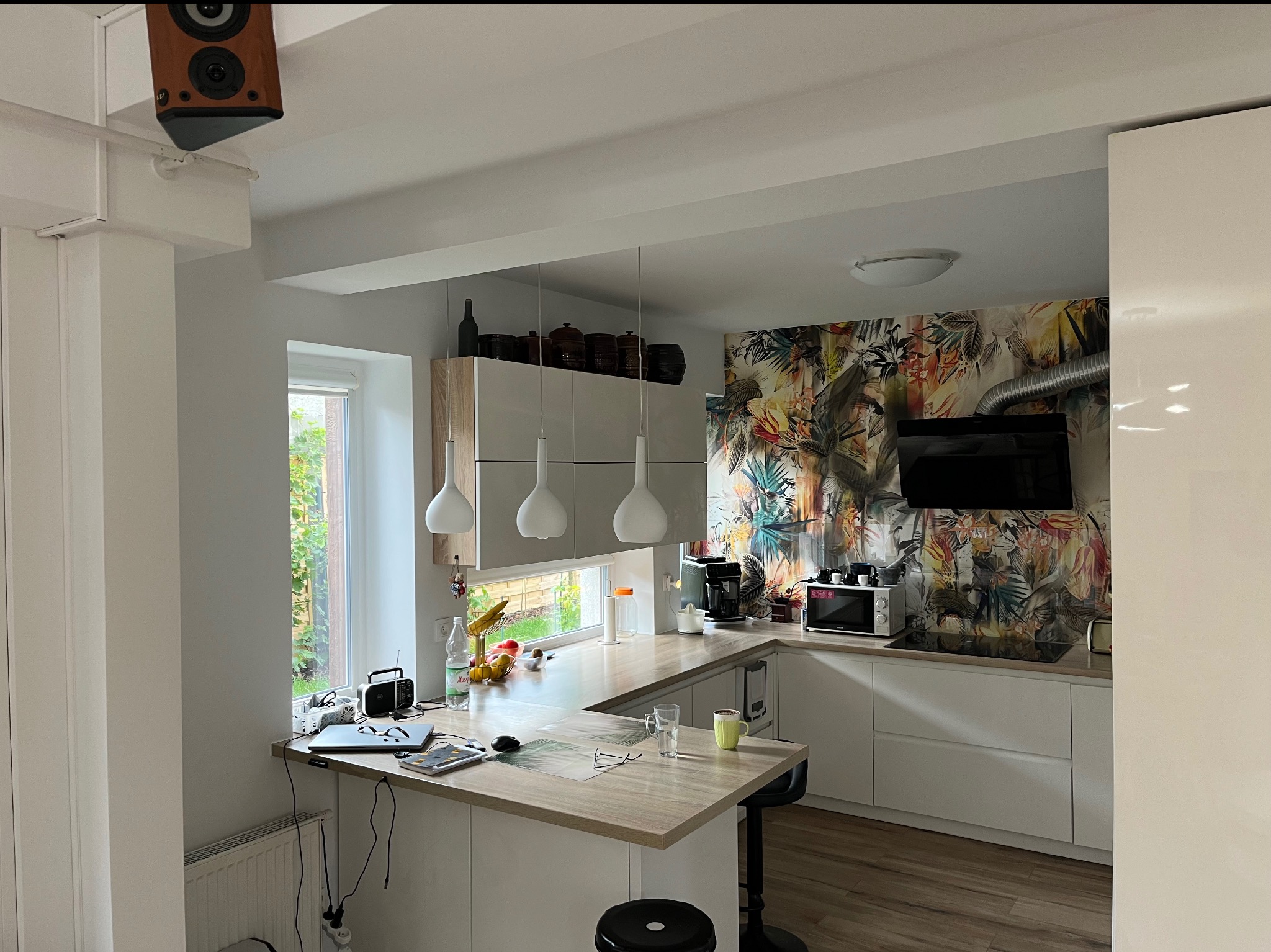 Nowoczesna kuchnia z wyspą, białymi szafkami i tropikalną tapetą. Widoczne owoce, laptop i głośnik na ścianie. Oświetlenie punktowe i okno z widokiem na zieleń.