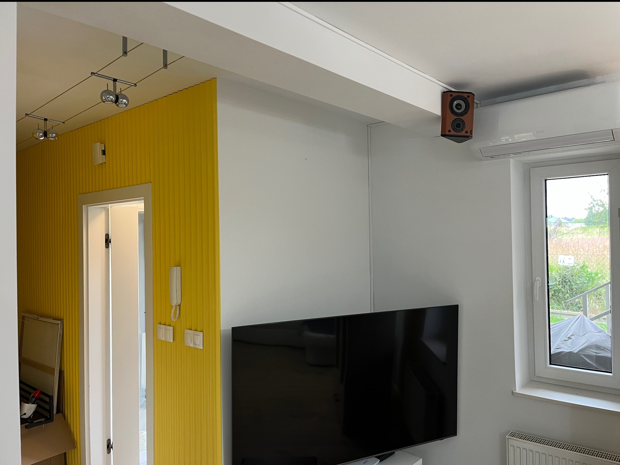 Żółta ściana z dekoracyjną fakturą w pokoju z białymi ścianami, oknem i telewizorem. Na suficie zamontowany głośnik i klimatyzator.