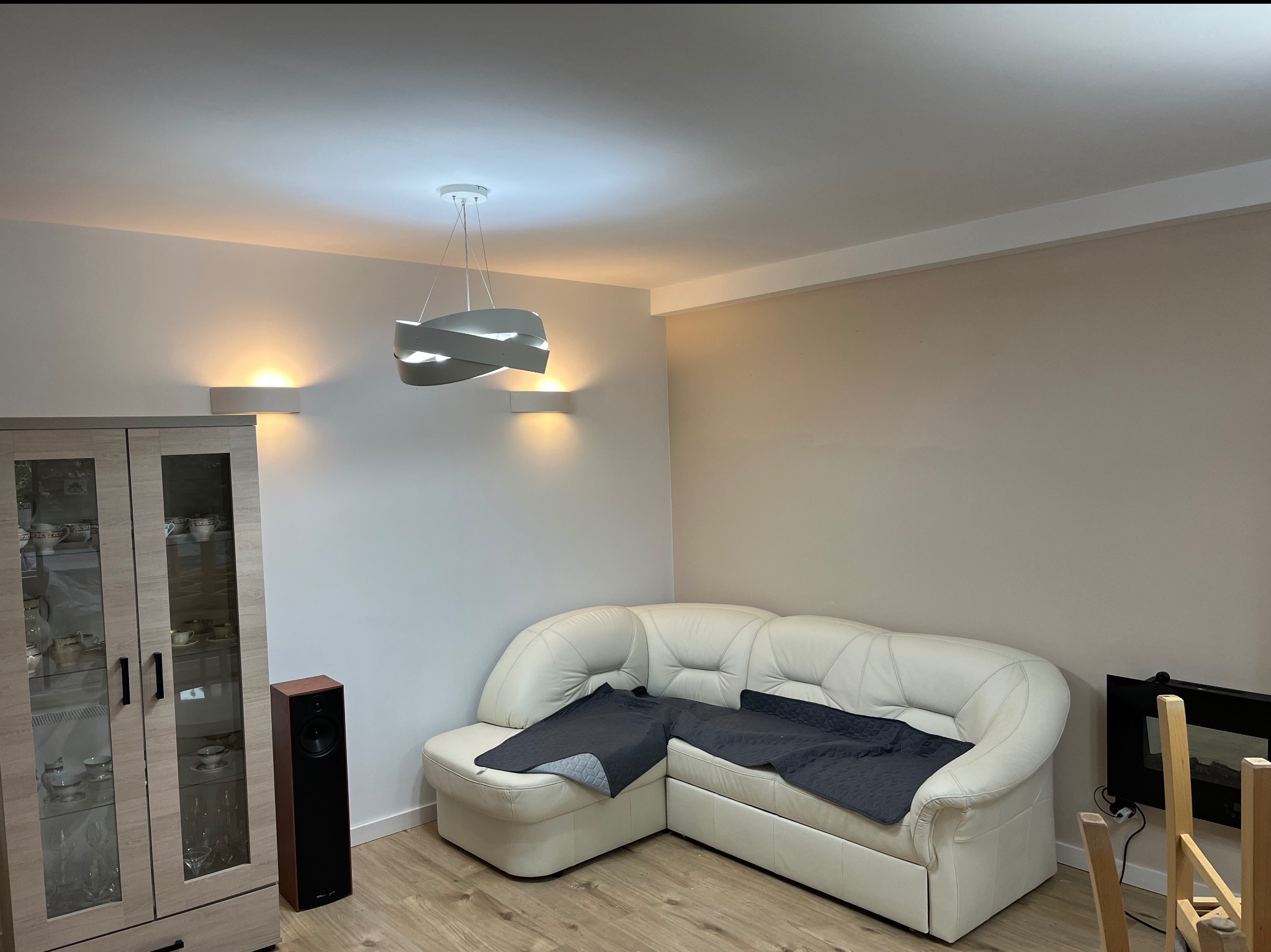 Jasne wnętrze salonu z narożną sofą, szafką z przeszkleniami i nowoczesnym oświetleniem. Ściany w neutralnych odcieniach, parkiet na podłodze.