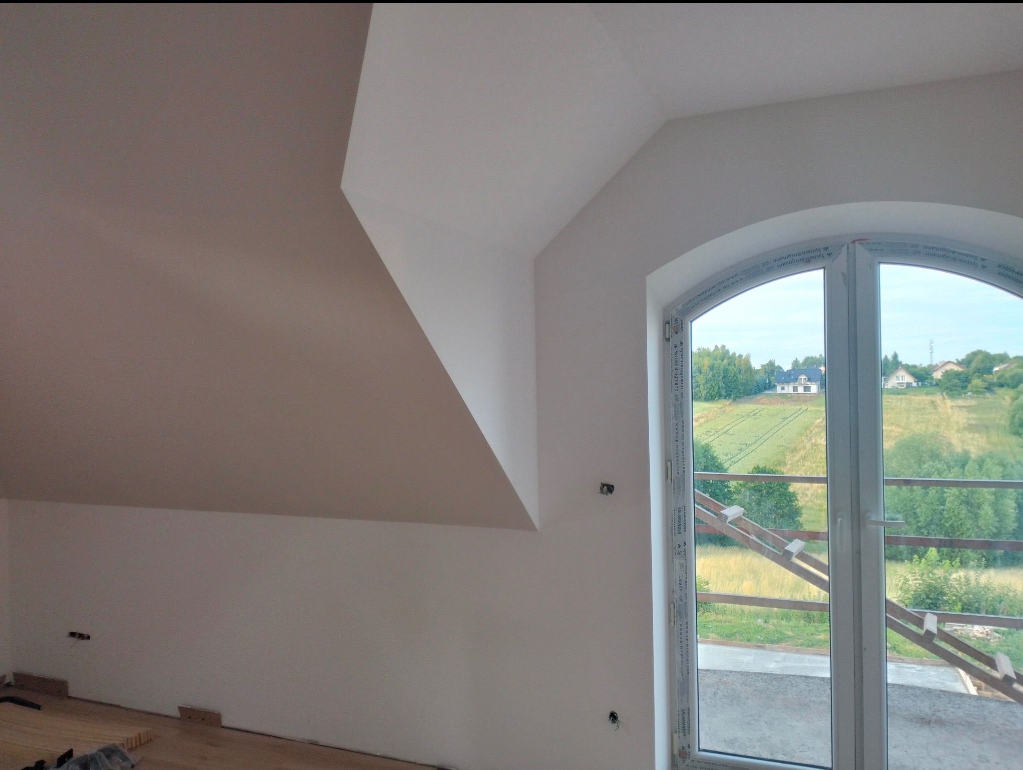 Wnętrze po malowaniu: białe ściany poddasza ze skosami i oknem łukowym z widokiem na zielone wzgórza. Częściowo widoczna podłoga w trakcie montażu.