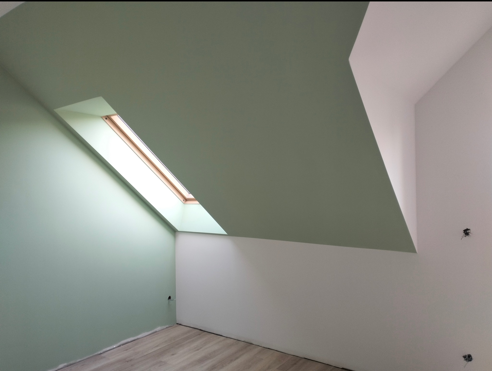 Pomalowane na zielono poddasze ze skośnym sufitem i oknem dachowym. Ściany częściowo białe, widoczne ślady po gniazdkach elektrycznych. Podłoga drewniana.