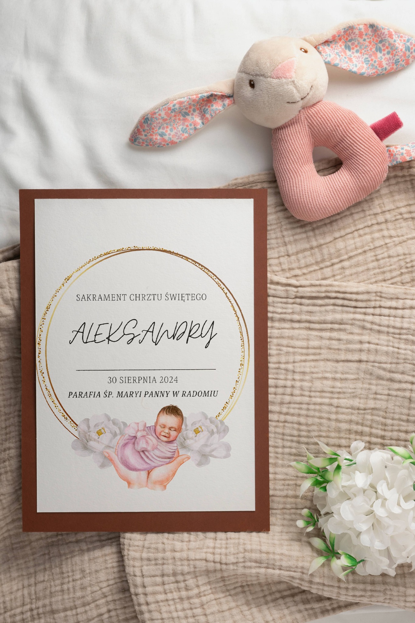 Zaproszenie na chrzest z imieniem Aleksandry, datą 30 sierpnia 2024 i ilustracją dziecka trzymanego w dłoniach, leży obok pluszowego króliczka i białych kwiatów na beżowym tle.