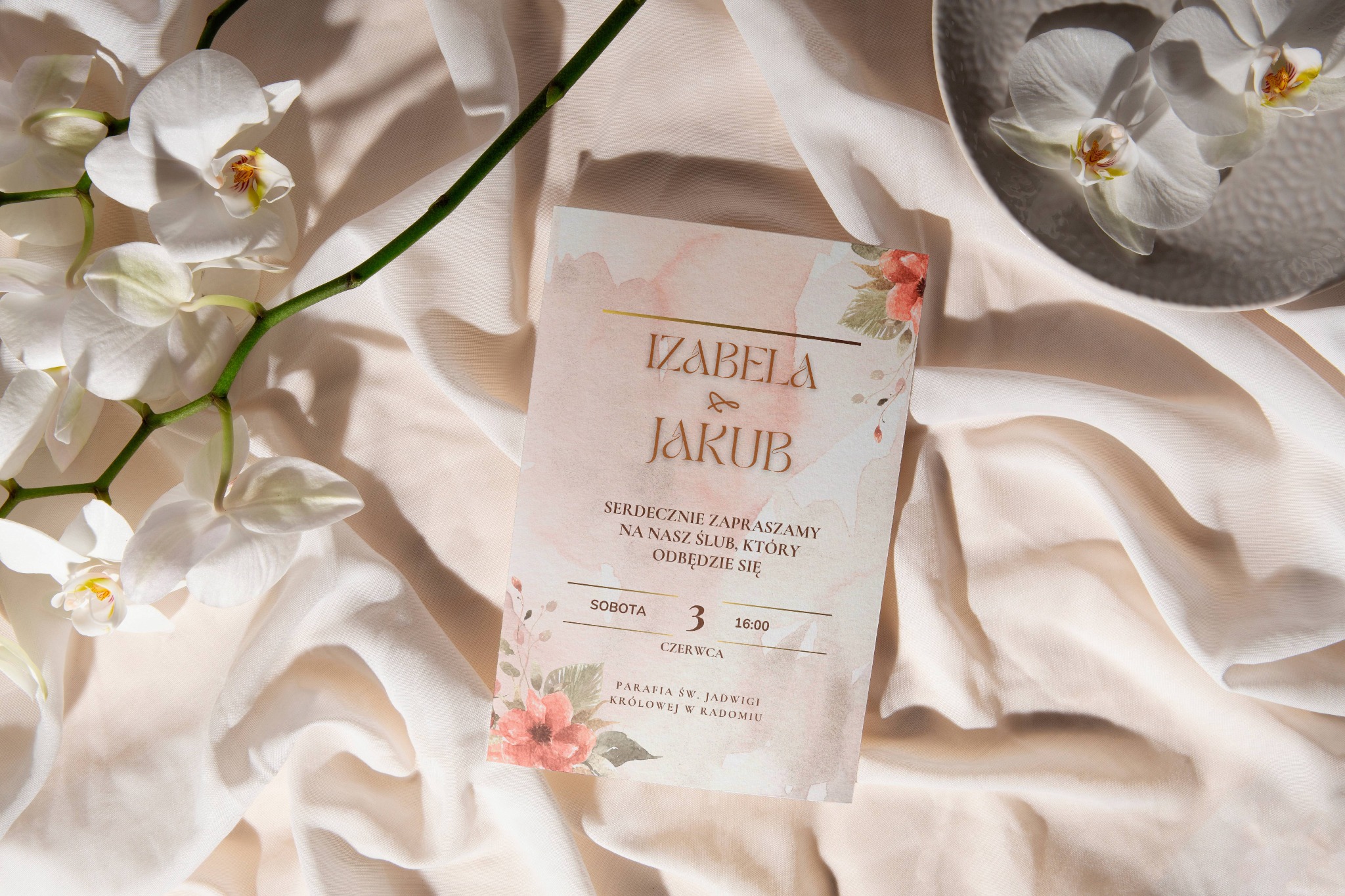 Eleganckie zaproszenie ślubne Izabeli i Jakuba z kwiatowym motywem, sfotografowane w Radomiu na tle kremowej tkaniny z orchideami, zapowiadające uroczystość w parafii św. Jadwigi Królowej.
