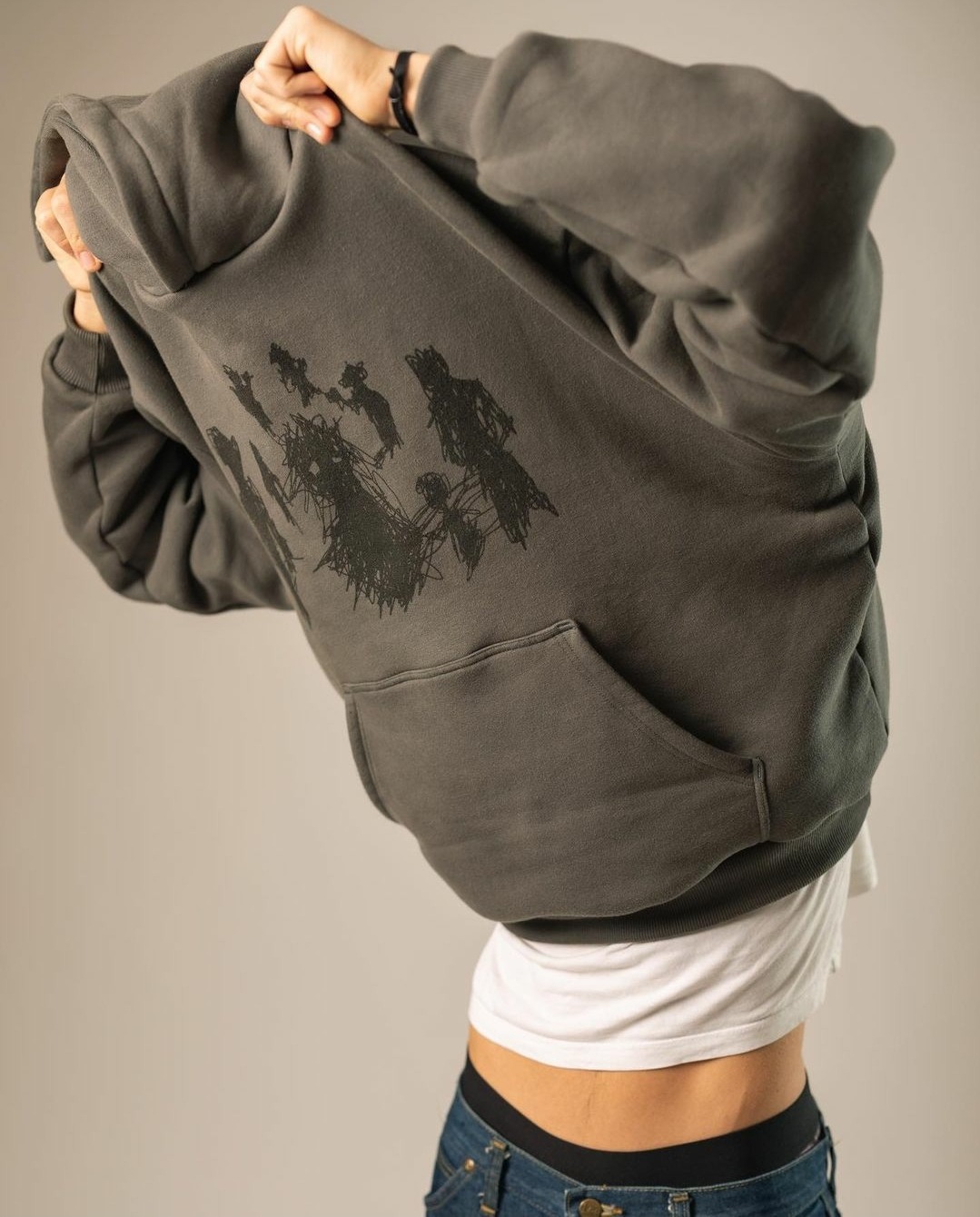 Osoba zdejmuje szarą bluzę z kapturem z abstrakcyjnym czarnym nadrukiem przedstawiającym postacie, widoczny fragment białej koszulki i dżinsów.