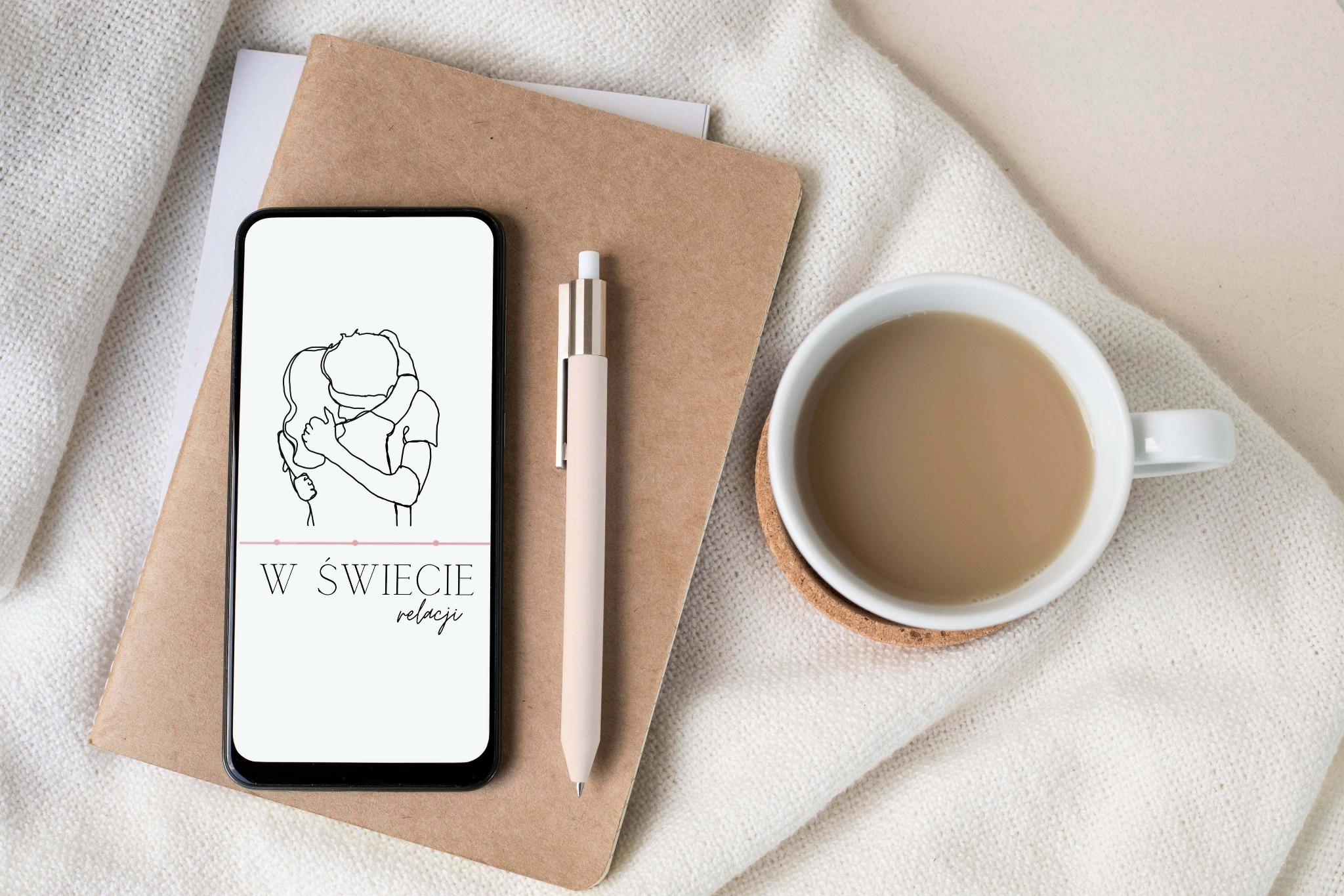 Smartfon z minimalistycznym rysunkiem obejmującej się pary i napisem 'W świecie relacji', leżący na brązowym notatniku obok beżowego długopisu i filiżanki kawy, na tle białej dzianiny.