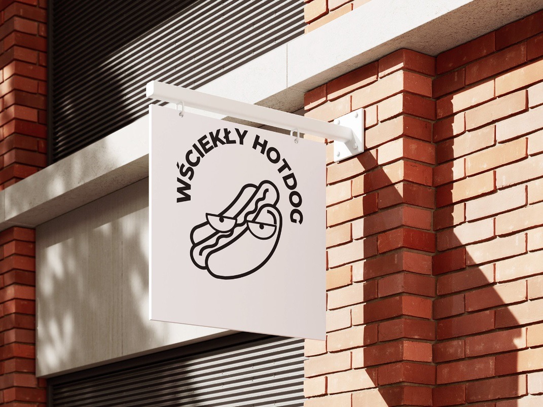 Szyld reklamowy z nazwą 'Wściekły Hotdog' i rysunkową grafiką hotdoga z okularami, zamontowany na ceglanej elewacji budynku.