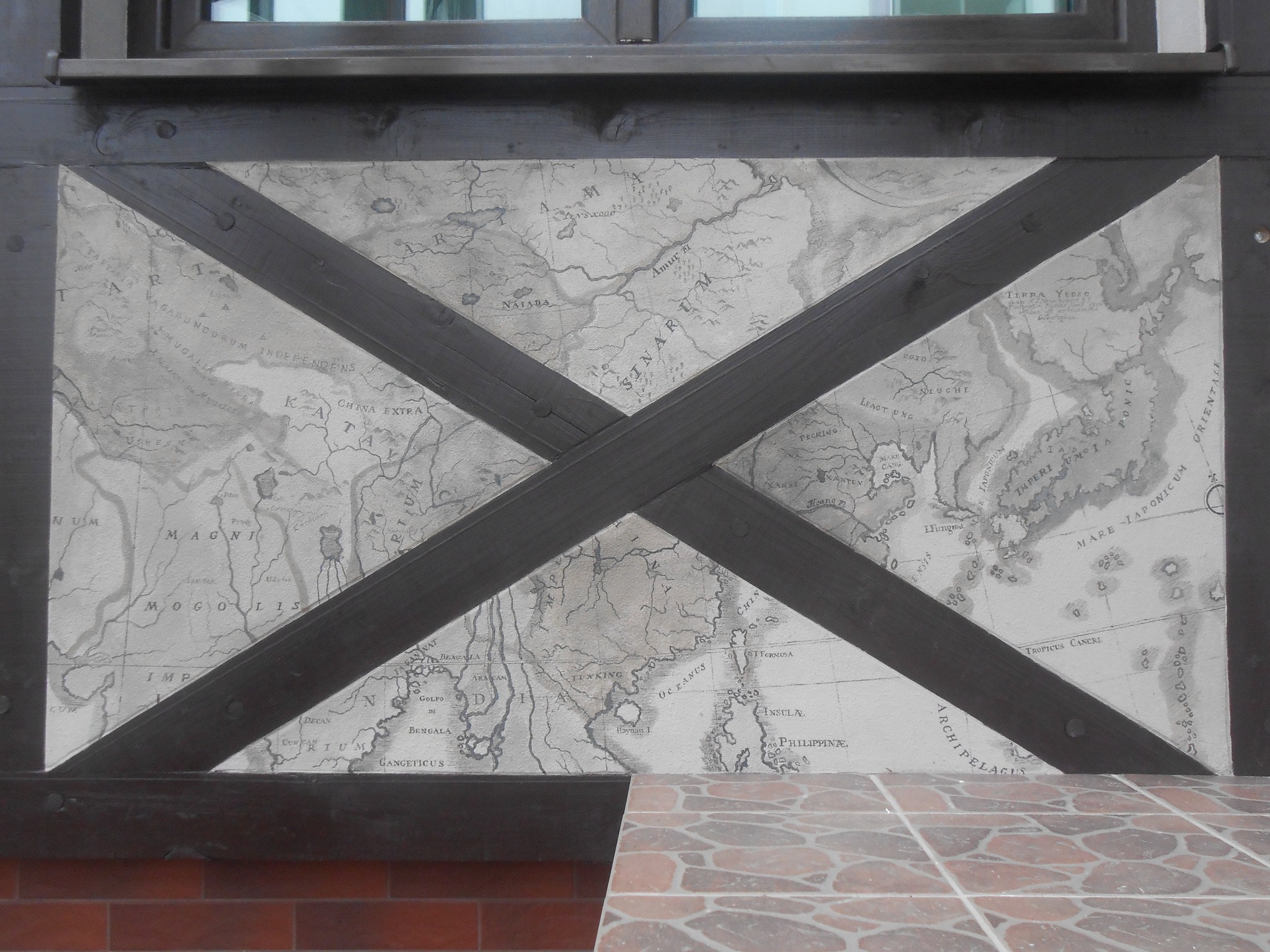 Fragment ściany z dekoracyjnym malowaniem przedstawiającym starą mapę Azji, przeciętej na skos drewnianymi belkami, widoczne fragmenty okna i płytek.