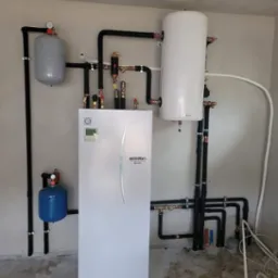 Biała pompa ciepła Ecodan Mitsubishi Electric z zbiornikami wyrównawczymi i czarnymi rurami w kotłowni, widoczne liczne zawory i manometry, na podłodze resztki po montażu.