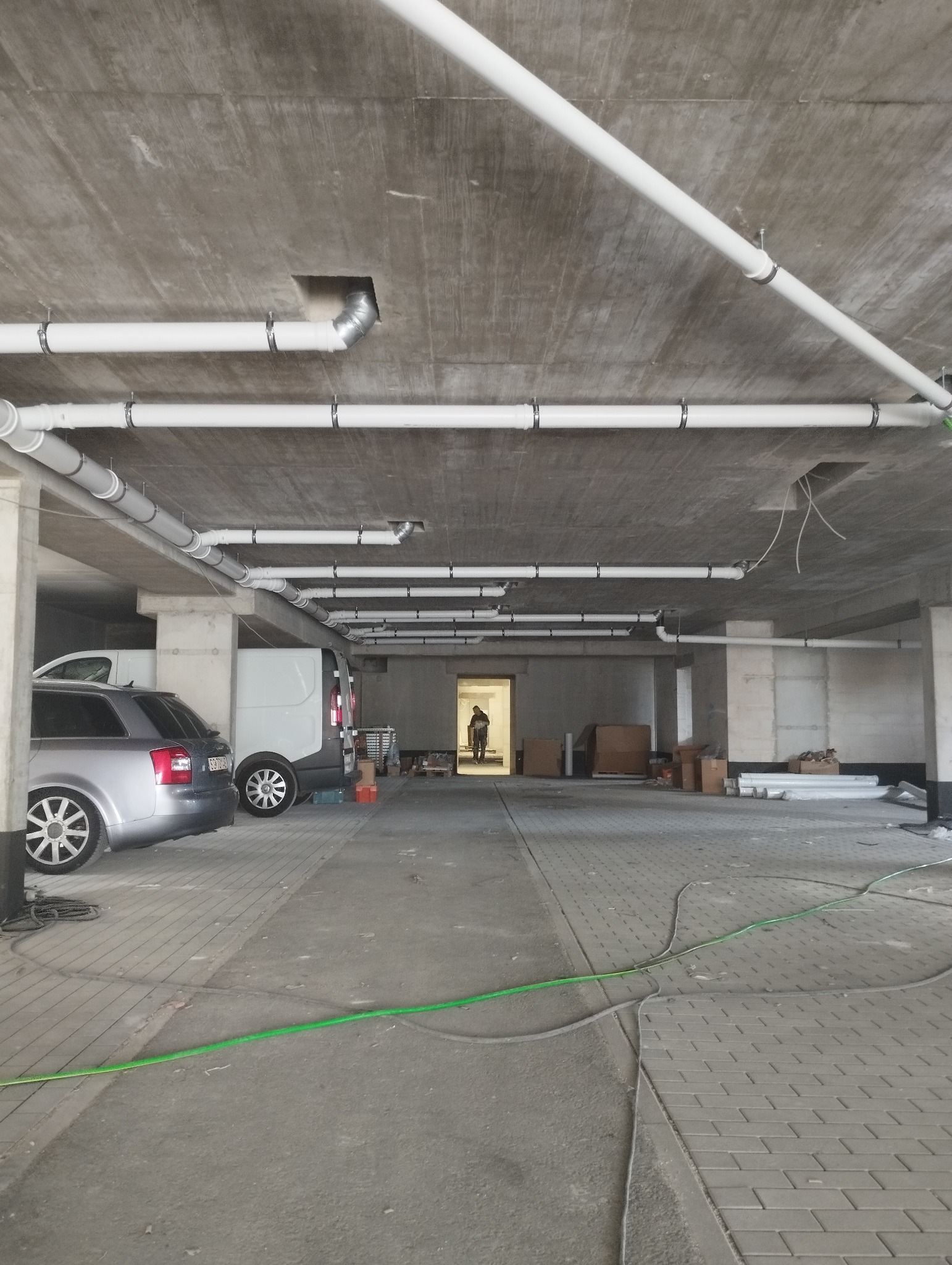 Instalacja białych rur pod sufitem parkingu podziemnego, widoczne samochody i osoba w oddali, przewody na podłodze.