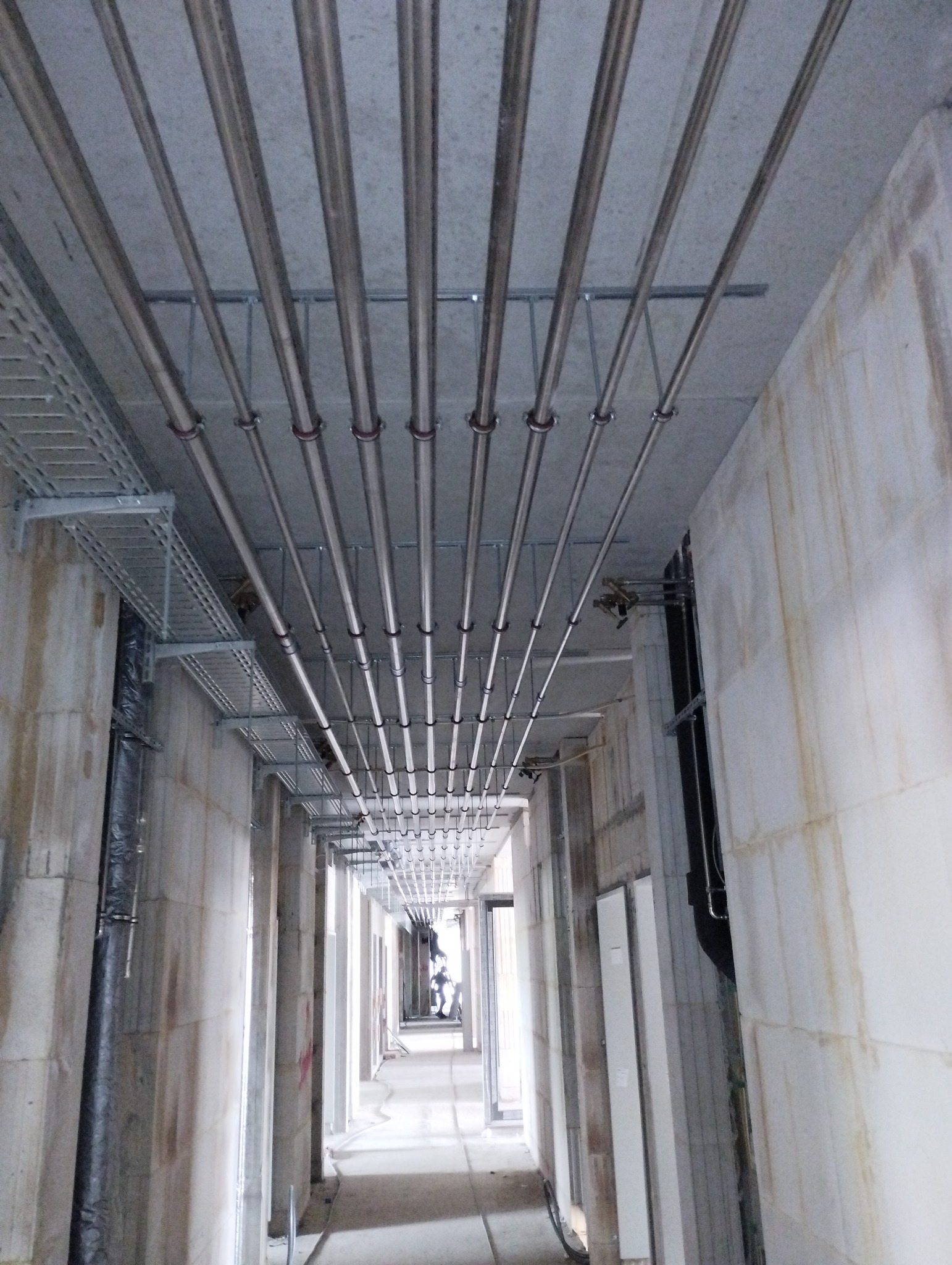 Wnętrze korytarza w surowym stanie z widocznymi metalowymi rurami instalacyjnymi pod sufitem, mocowanymi za pomocą metalowych uchwytów i szyn. Dodatkowo widoczne metalowe korytka kablowe na ścianach.