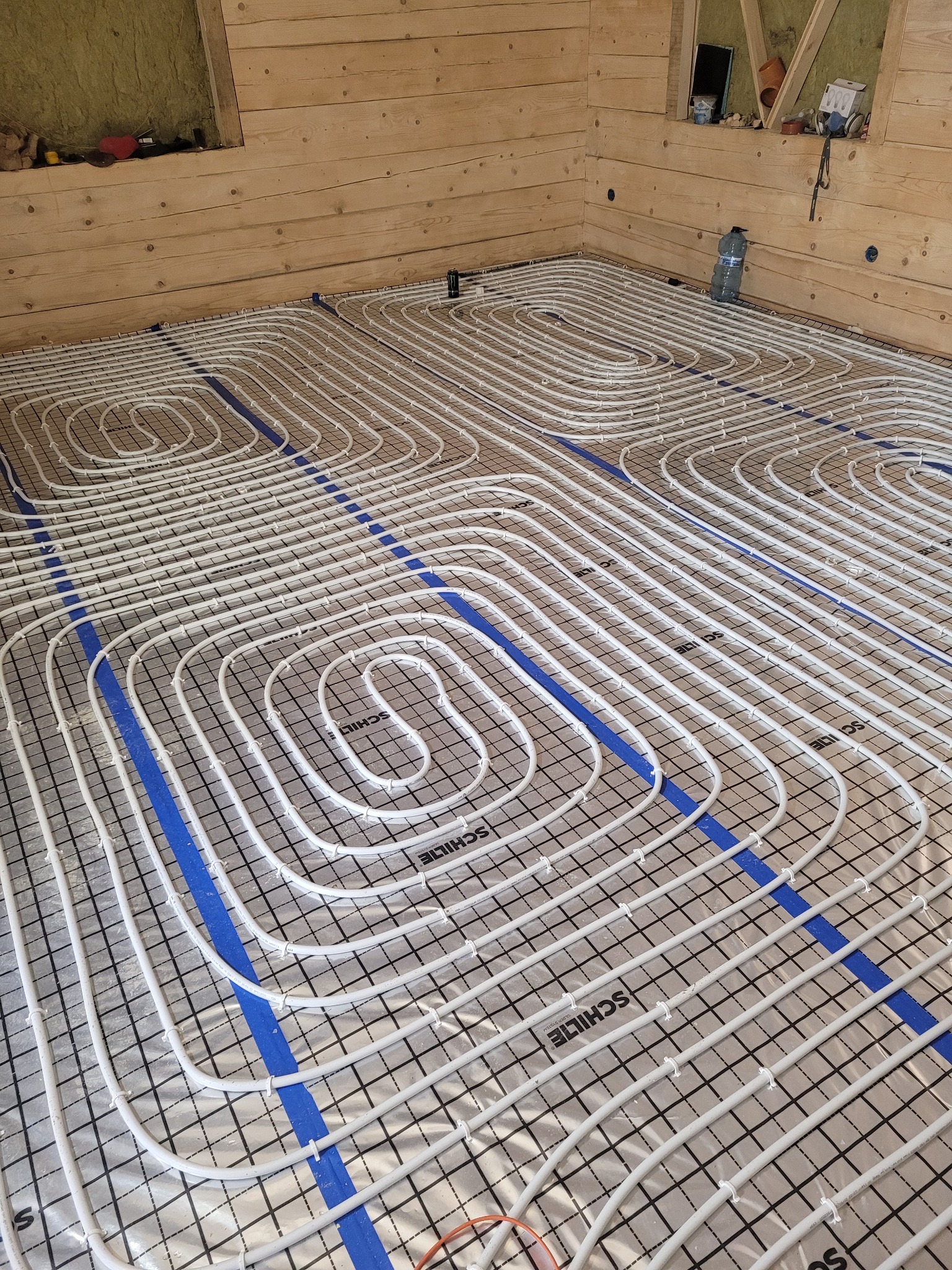Białe rurki ogrzewania podłogowego ułożone w pętle na srebrnej folii z czarną siatką, przymocowane niebieską taśmą, widoczne drewniane ściany i poddasze