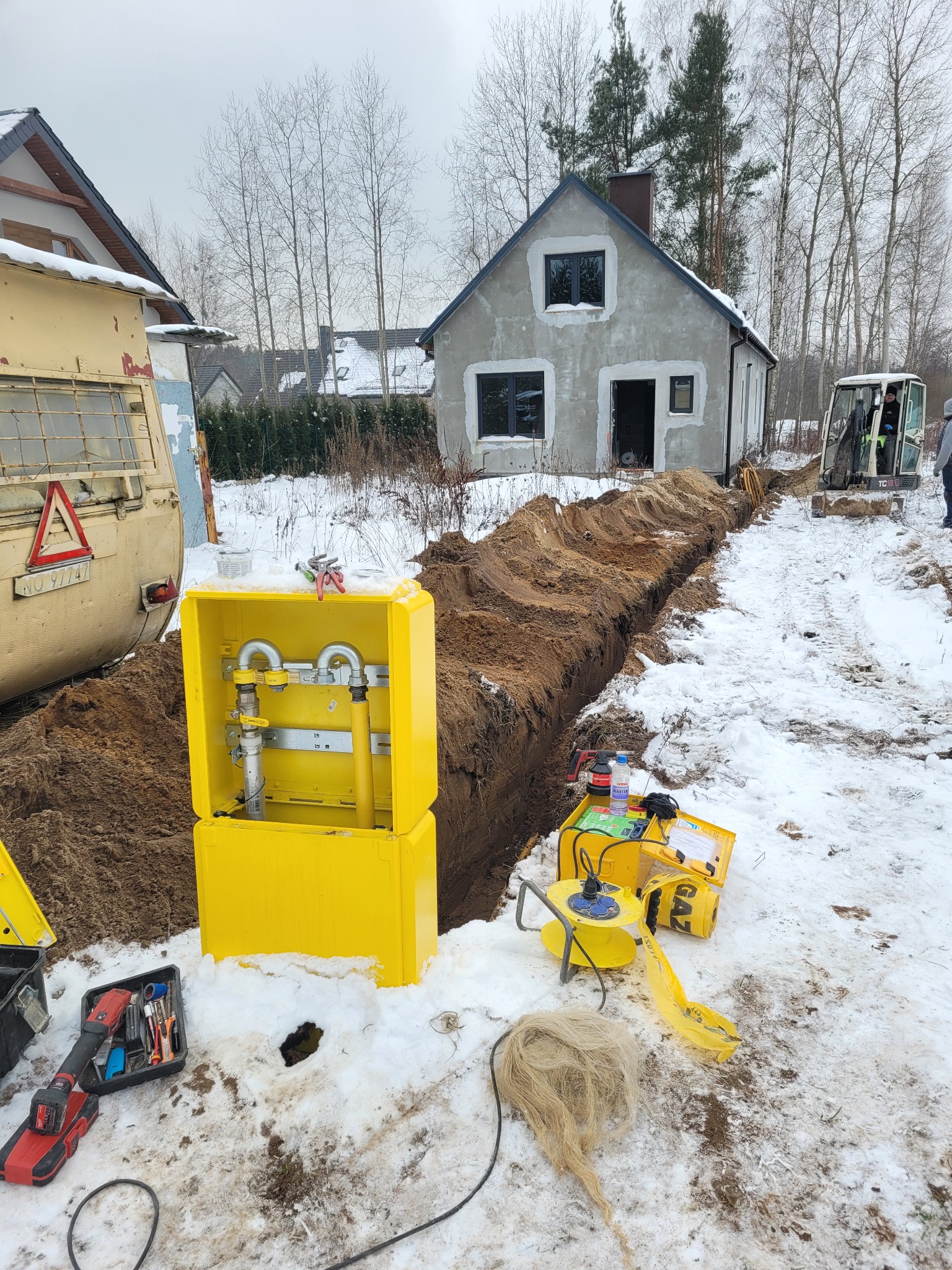 Żółta skrzynka gazowa z instalacją, narzędzia i wykop w zasypanym śniegiem terenie budowy domu jednorodzinnego z mini koparką w tle.