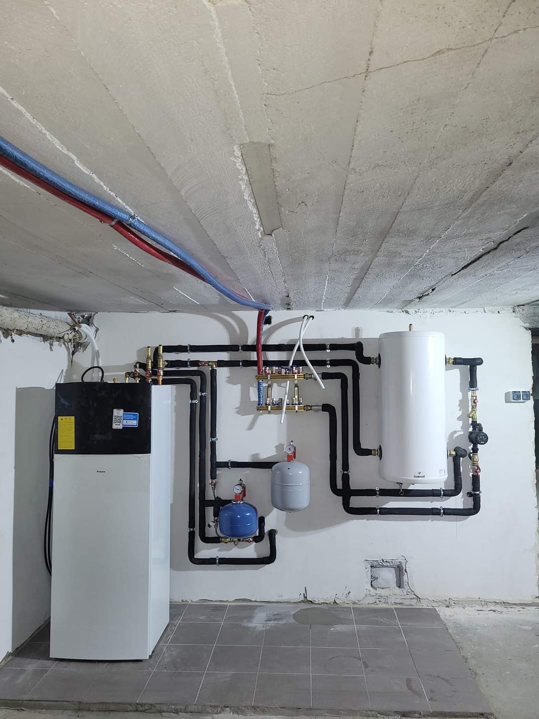 Pompa ciepła Daikin, zbiornik akumulacyjny, naczynia przeponowe i instalacja hydrauliczna z czarnych rur w piwnicy z surowym betonowym sufitem i szarą podłogą z płytek.