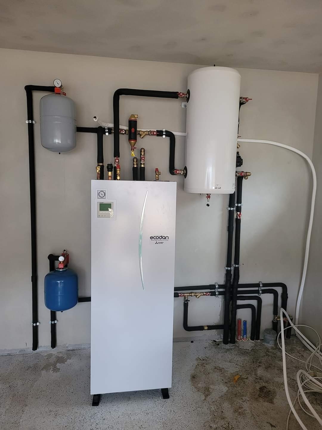 Biała pompa ciepła Ecodan Mitsubishi Electric z zbiornikami wyrównawczymi i czarnymi rurami w kotłowni, widoczne liczne zawory i manometry, na podłodze resztki po montażu.