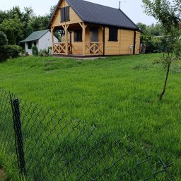 FHU EARTHWORKS AND GARDENING - Drewniany domek letniskowy z czarnym dachem na zielonym trawniku, ogrodzony siatką. W tle widoczne inne budynki i drzewa.