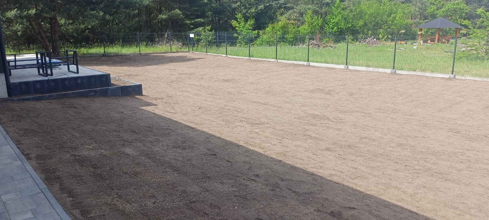 Przygotowany teren pod trawnik z widoczną ziemią wyrównaną i ogrodzeniem w tle, częściowo zacieniony.