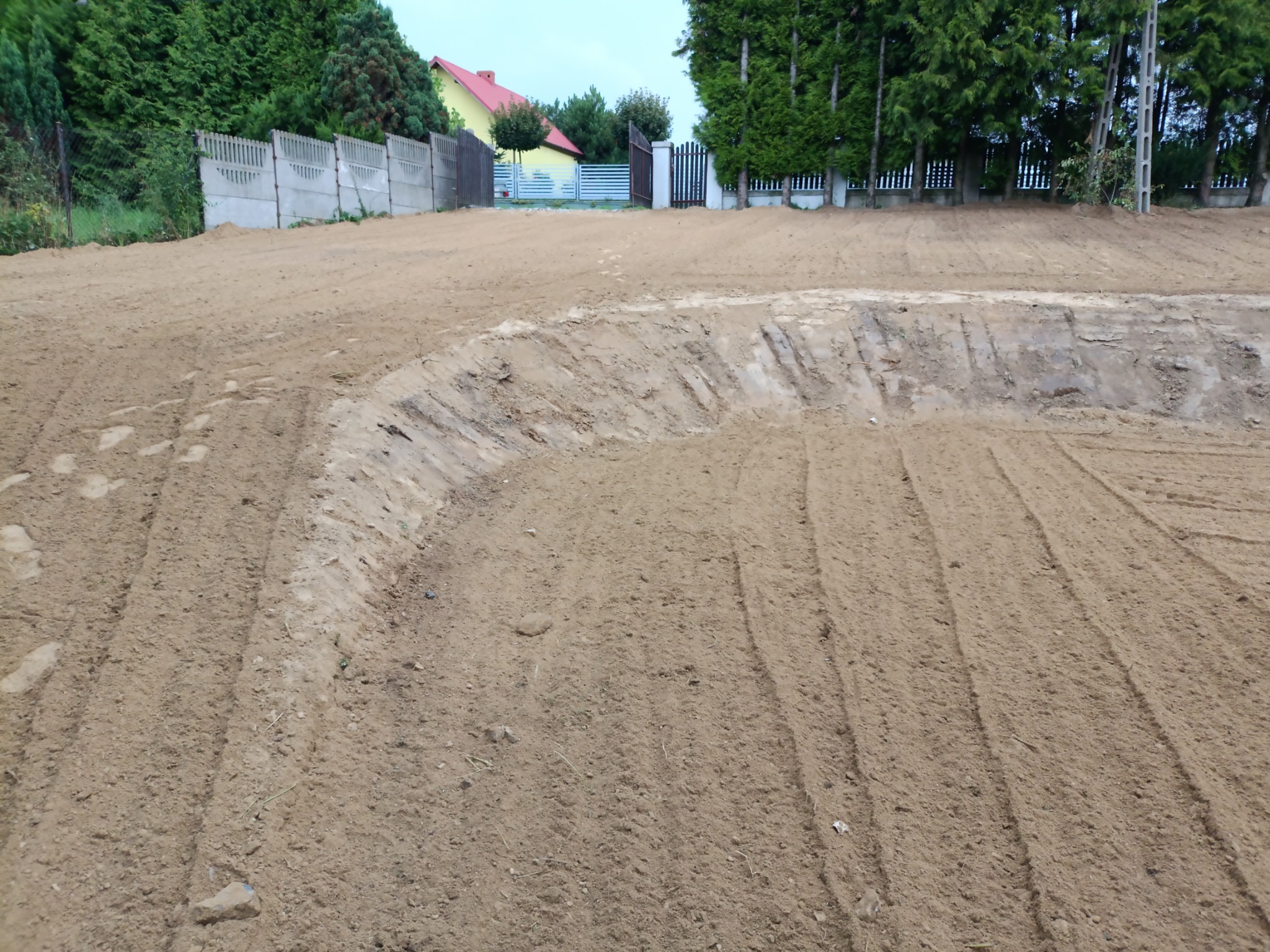 Przygotowany teren pod trawnik z widocznymi śladami bronowania, w tle ogrodzenie i zabudowania.