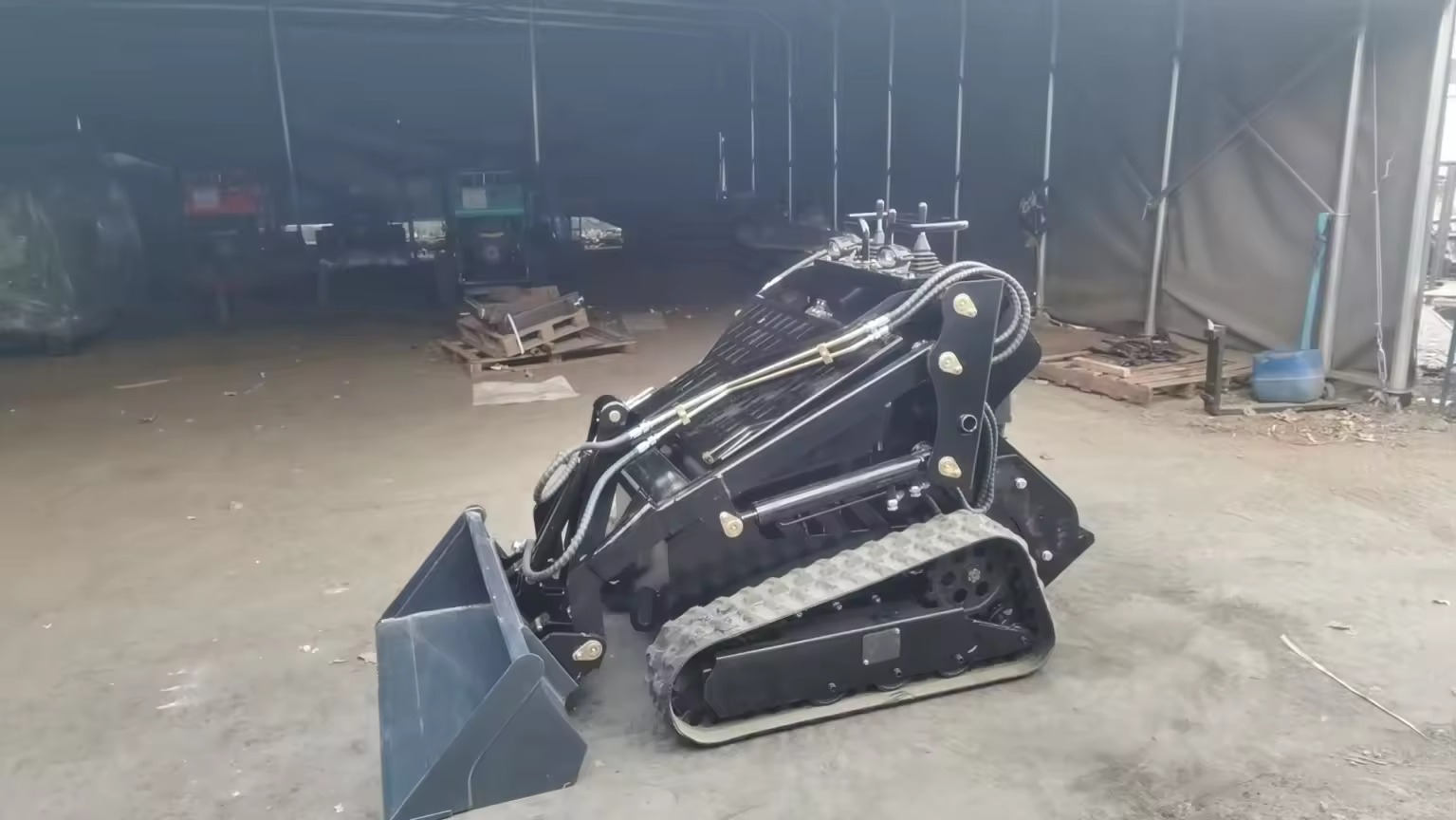 Czarna miniładowarka gąsienicowa z łyżką, stojąca w hali magazynowej, widok z boku, widoczne detale konstrukcyjne i hydraulika