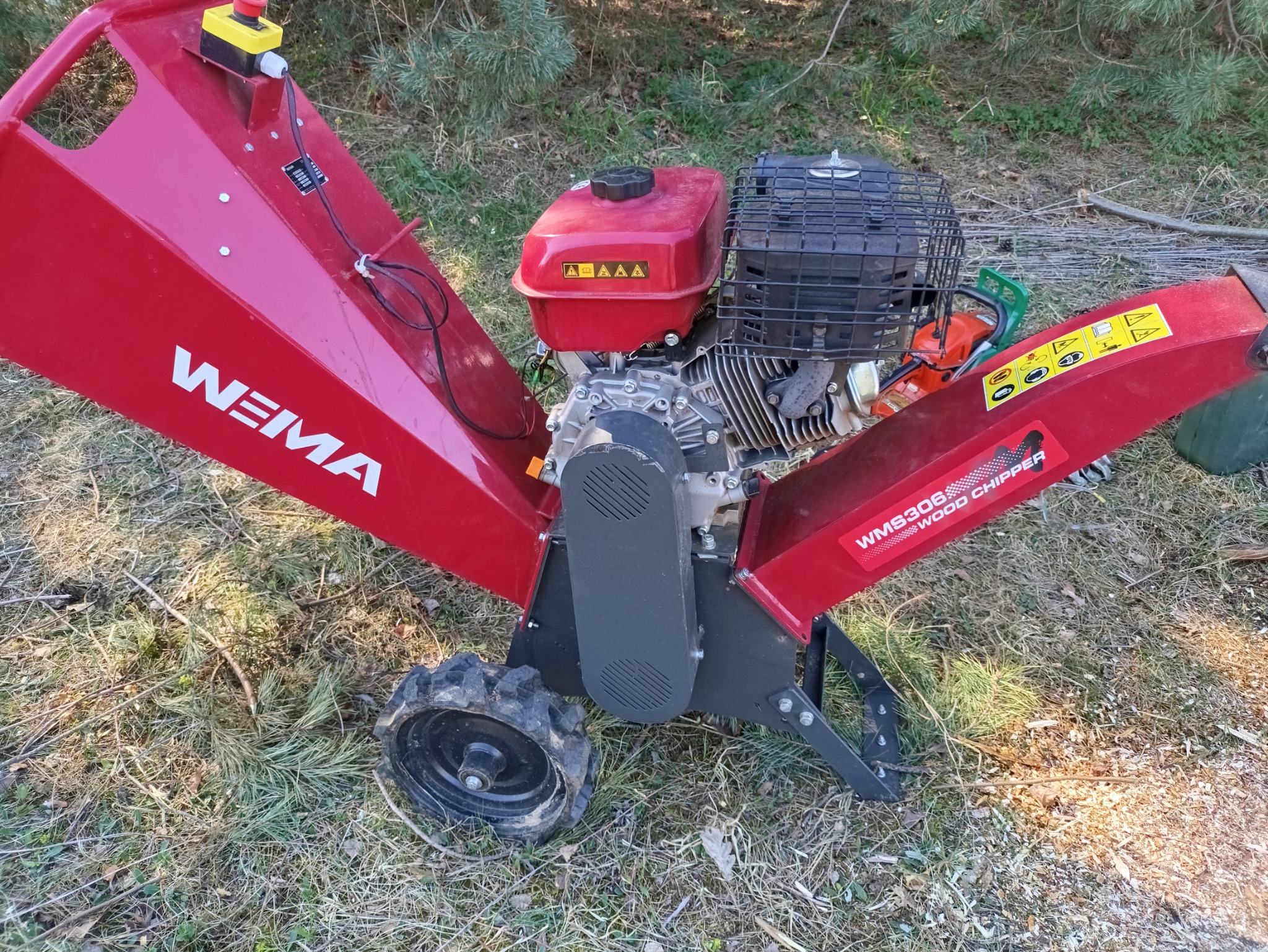 Czerwony, spalinowy rębak do gałęzi marki Weima, model WMS306, stojący na trawie z widoczną obok ściętą gałęzią.