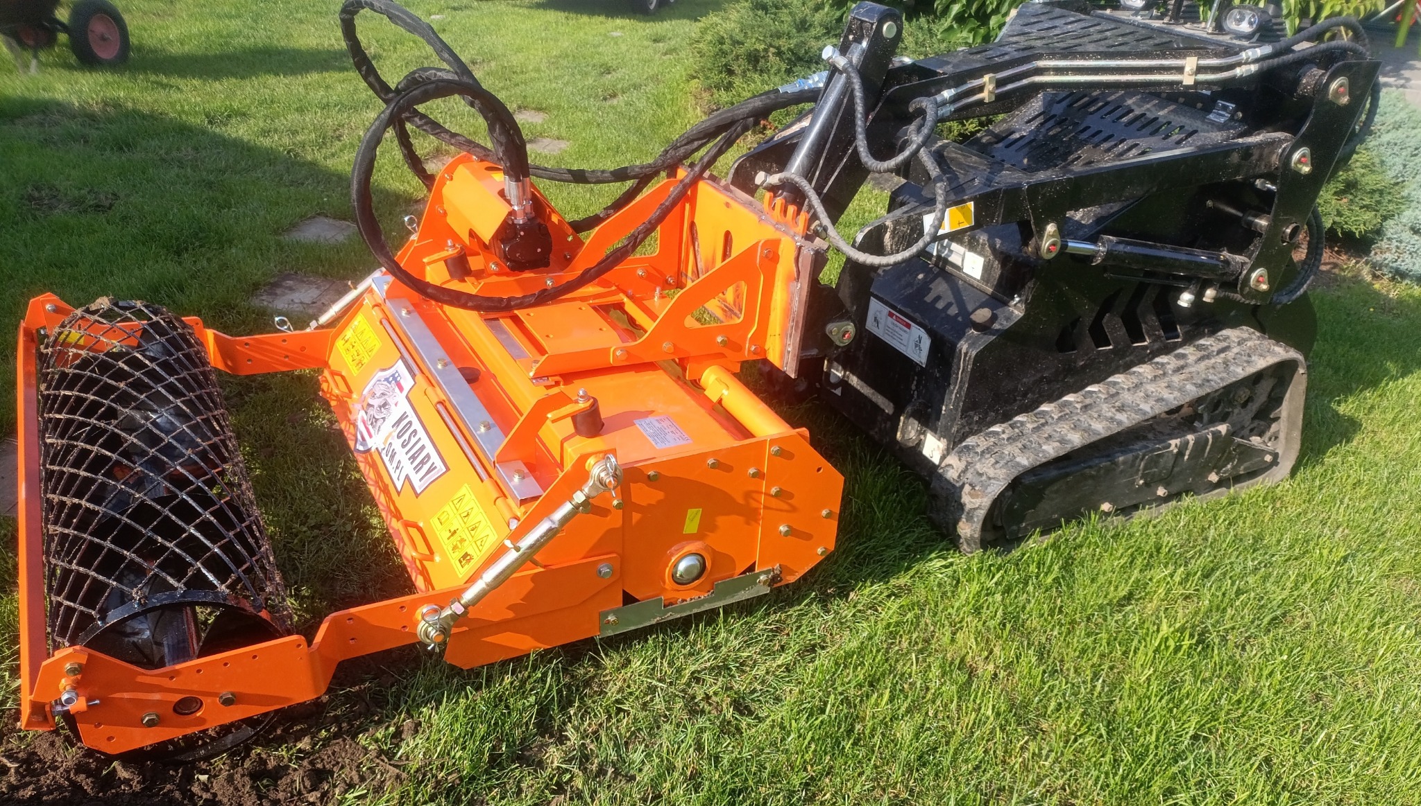 Czarny miniładowarka gąsienicowa z pomarańczowym frezem glebowym Kosiary, przygotowująca teren pod trawnik na zielonej trawie w słoneczny dzień.