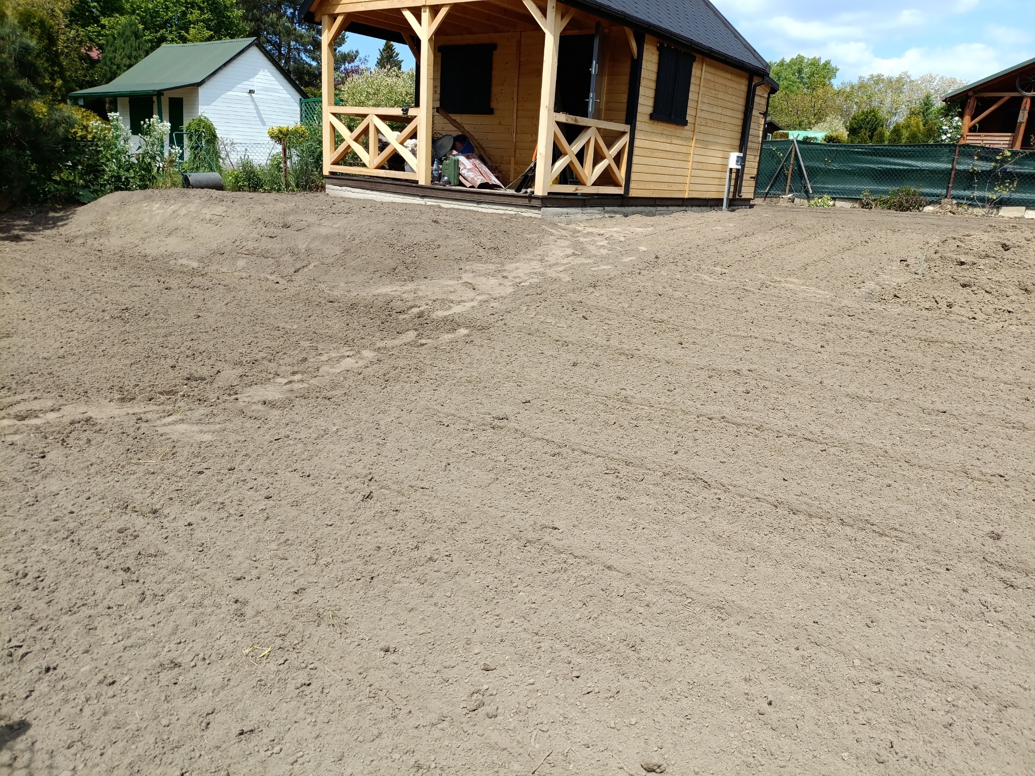 Przygotowany teren pod trawnik z widocznymi śladami po maszynach, obok drewniany domek letniskowy i zielone ogrodzenie z siatki.