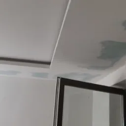 Niedokończone szpachlowanie sufitu z widocznymi łatami świeżo nałożonej masy szpachlowej, widok z dołu na fragment ściany i okno.