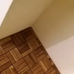 Fragment podłogi wyłożonej panelami drewnianymi ułożonymi w wzór jodełki, widoczny narożnik pomieszczenia z białymi ścianami.