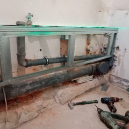 Krzysztof Wojciechowski - Stelaż pod zabudowę WC z widocznymi rurami kanalizacyjnymi, poziomowany laserowo. Na podłodze narzędzia: młotek, wiertarki, poziomica. Surowy stan pomieszczenia.