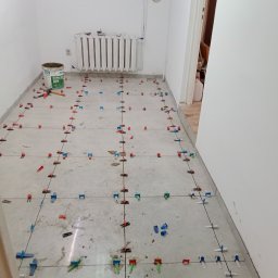 Krzysztof Wojciechowski - Szare płytki podłogowe w trakcie układania, widoczne systemy poziomowania płytek (klipsy i kliny) w kolorach: czerwonym, niebieskim i zielonym. Wiadro z zaprawą obok grzejnika.