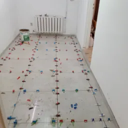 Szare płytki podłogowe w trakcie układania, widoczne systemy poziomowania płytek (klipsy i kliny) w kolorach: czerwonym, niebieskim i zielonym. Wiadro z zaprawą obok grzejnika.