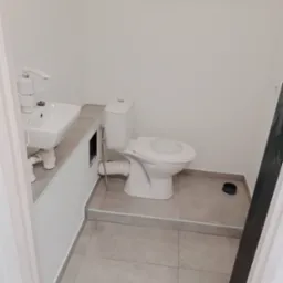 Biała umywalka z baterią i toaleta w małej łazience, szare płytki podłogowe. Widoczne rury i odpływ. Minimalistyczne wykończenie.