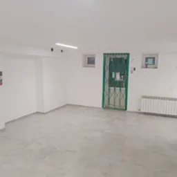 Białe, odświeżone wnętrze z szarą podłogą z płytek. Zielona krata w oknie i biały grzejnik. Minimalistyczne oświetlenie sufitowe.