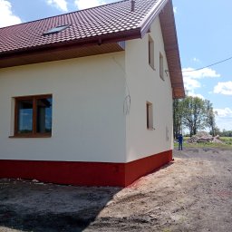 Krzysztof Wojciechowski - Świeżo otynkowany, dwukondygnacyjny dom z brązowym dachem i oknami, z czerwonym cokołem, na tle błękitnego nieba i zieleni. Widoczne niedociągnięcia.