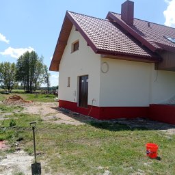 Krzysztof Wojciechowski - Świeżo pomalowany dom z czerwoną podmurówką i dachem z dachówki w słoneczny dzień. Widoczne narzędzia malarskie i krajobraz w tle.