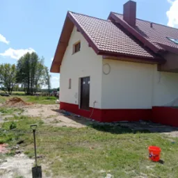 Świeżo pomalowany dom z czerwoną podmurówką i dachem z dachówki w słoneczny dzień. Widoczne narzędzia malarskie i krajobraz w tle.