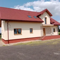 Elewacja domu jednorodzinnego z czerwonym dachem, kremową fasadą i kamiennym wykończeniem wejścia. Widoczne okna z drewnianą ramą i fragment otoczenia.