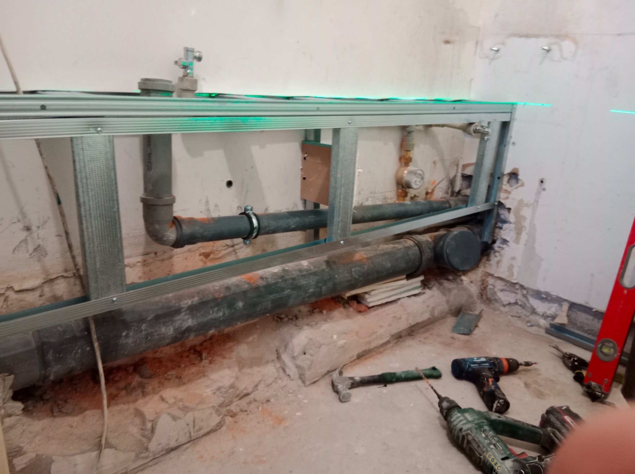 Stelaż pod zabudowę WC z widocznymi rurami kanalizacyjnymi, poziomowany laserowo. Na podłodze narzędzia: młotek, wiertarki, poziomica. Surowy stan pomieszczenia.