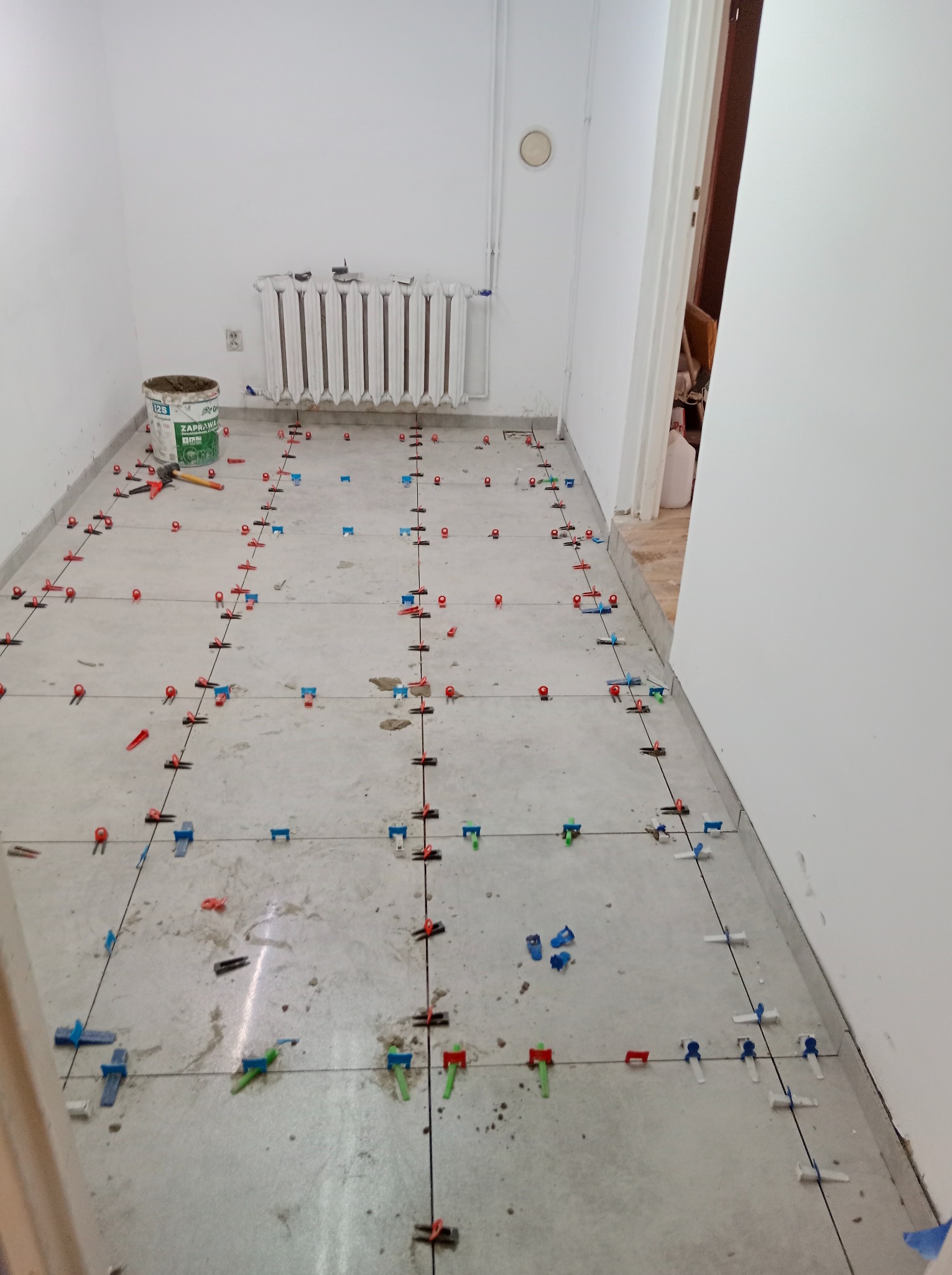 Szare płytki podłogowe w trakcie układania, widoczne systemy poziomowania płytek (klipsy i kliny) w kolorach: czerwonym, niebieskim i zielonym. Wiadro z zaprawą obok grzejnika.