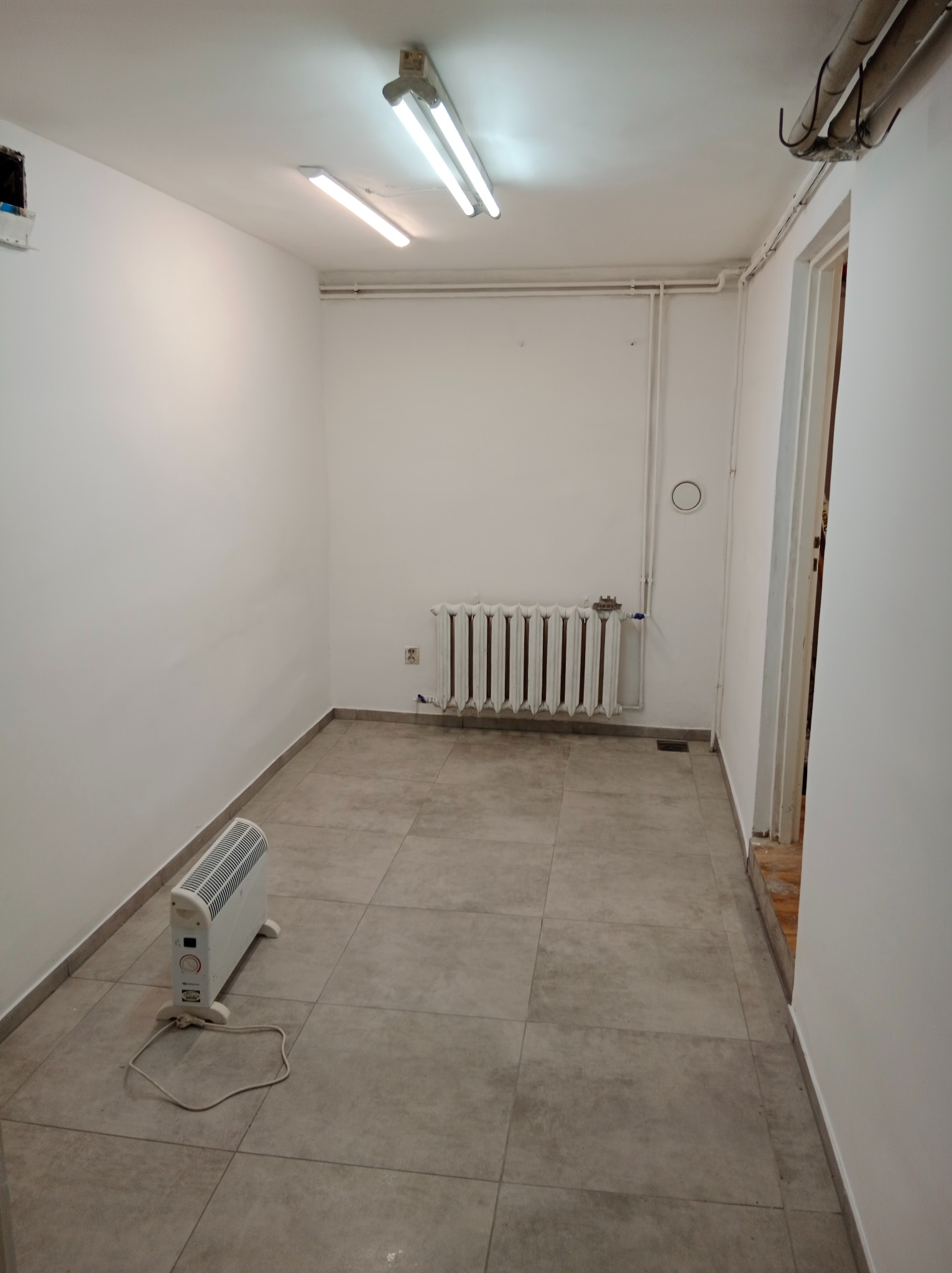 Puste, odświeżone wnętrze pokoju z szarą podłogą, białymi ścianami, grzejnikiem i przenośnym grzejnikiem elektrycznym. Widoczne rury pod sufitem i oświetlenie.