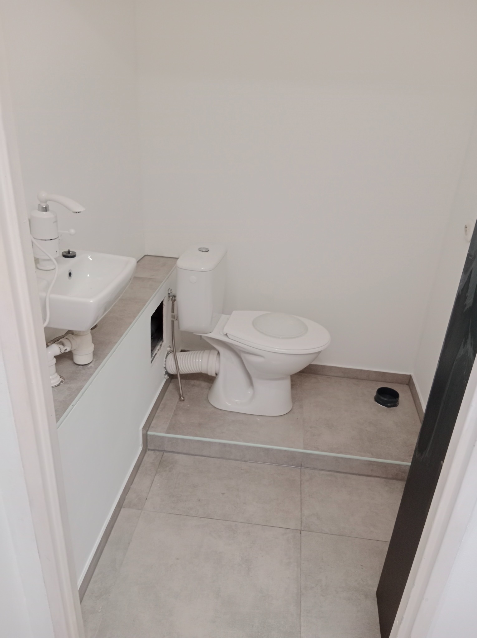 Biała umywalka z baterią i toaleta w małej łazience, szare płytki podłogowe. Widoczne rury i odpływ. Minimalistyczne wykończenie.