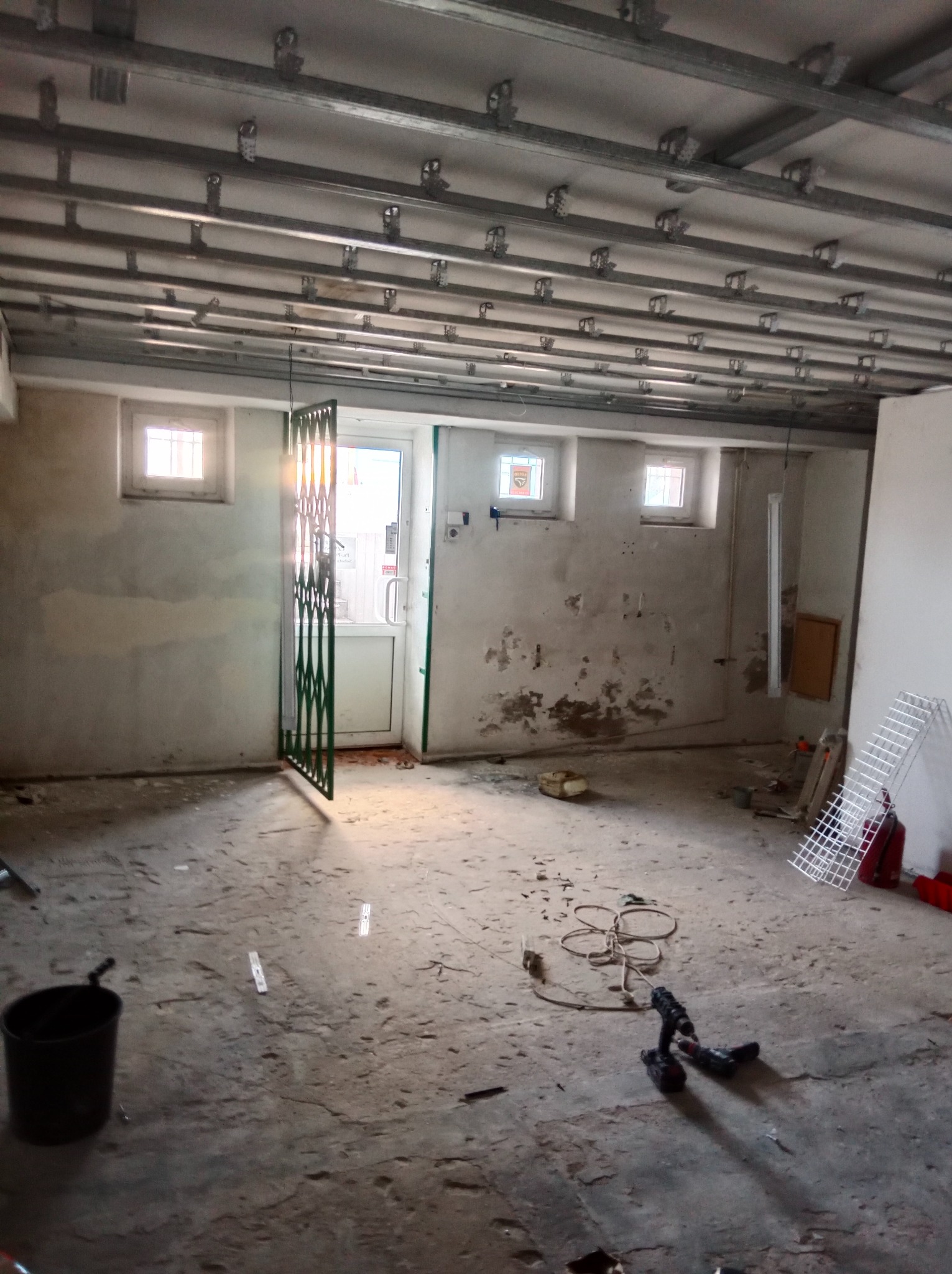 Metalowa konstrukcja sufitu podwieszanego z płyt karton-gips, widok na surowy betonowy podkład, narzędzia pracy i okno z kratą w tle. Etap prac wykończeniowych.