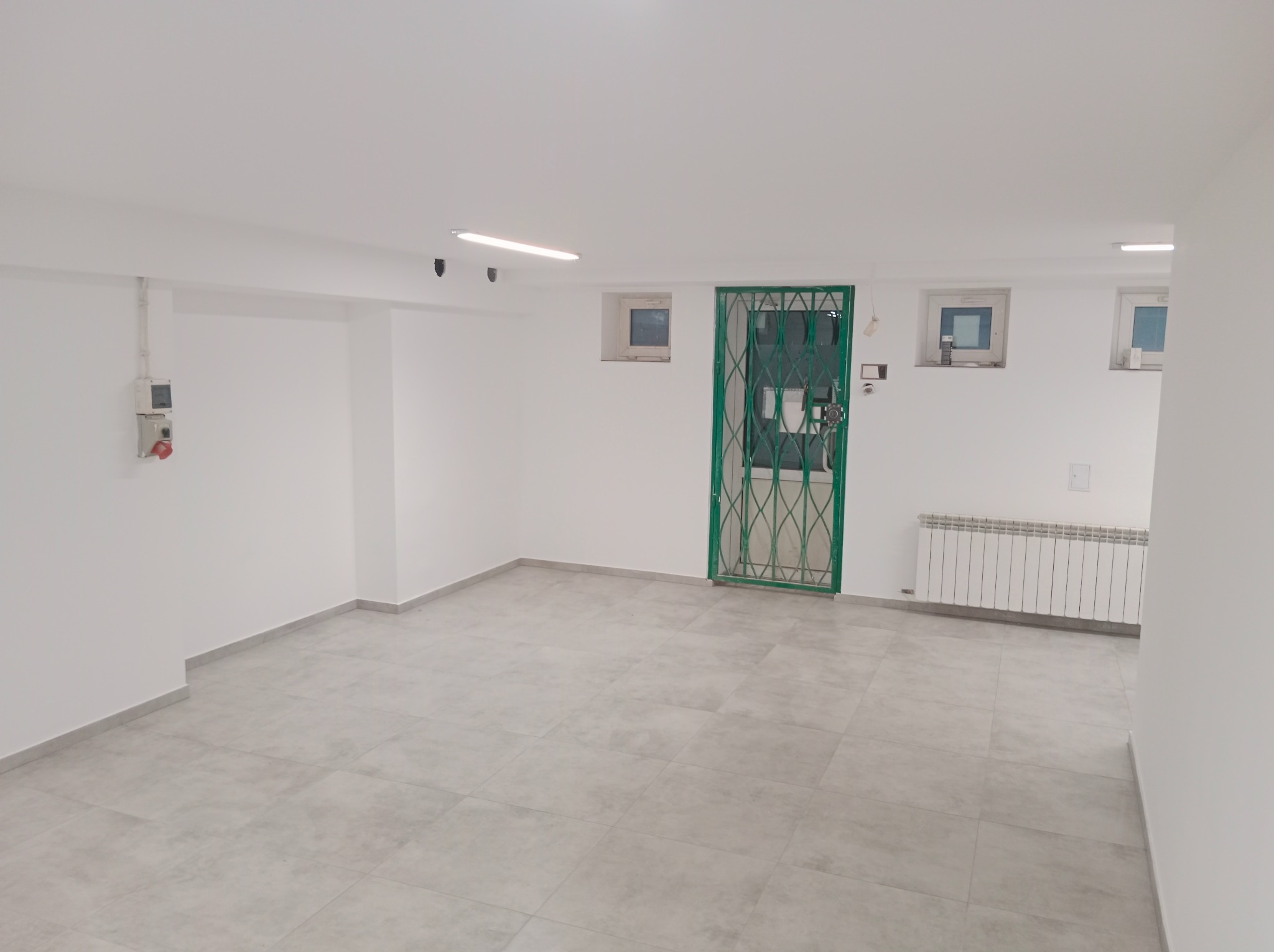 Białe, odświeżone wnętrze z szarą podłogą z płytek. Zielona krata w oknie i biały grzejnik. Minimalistyczne oświetlenie sufitowe.