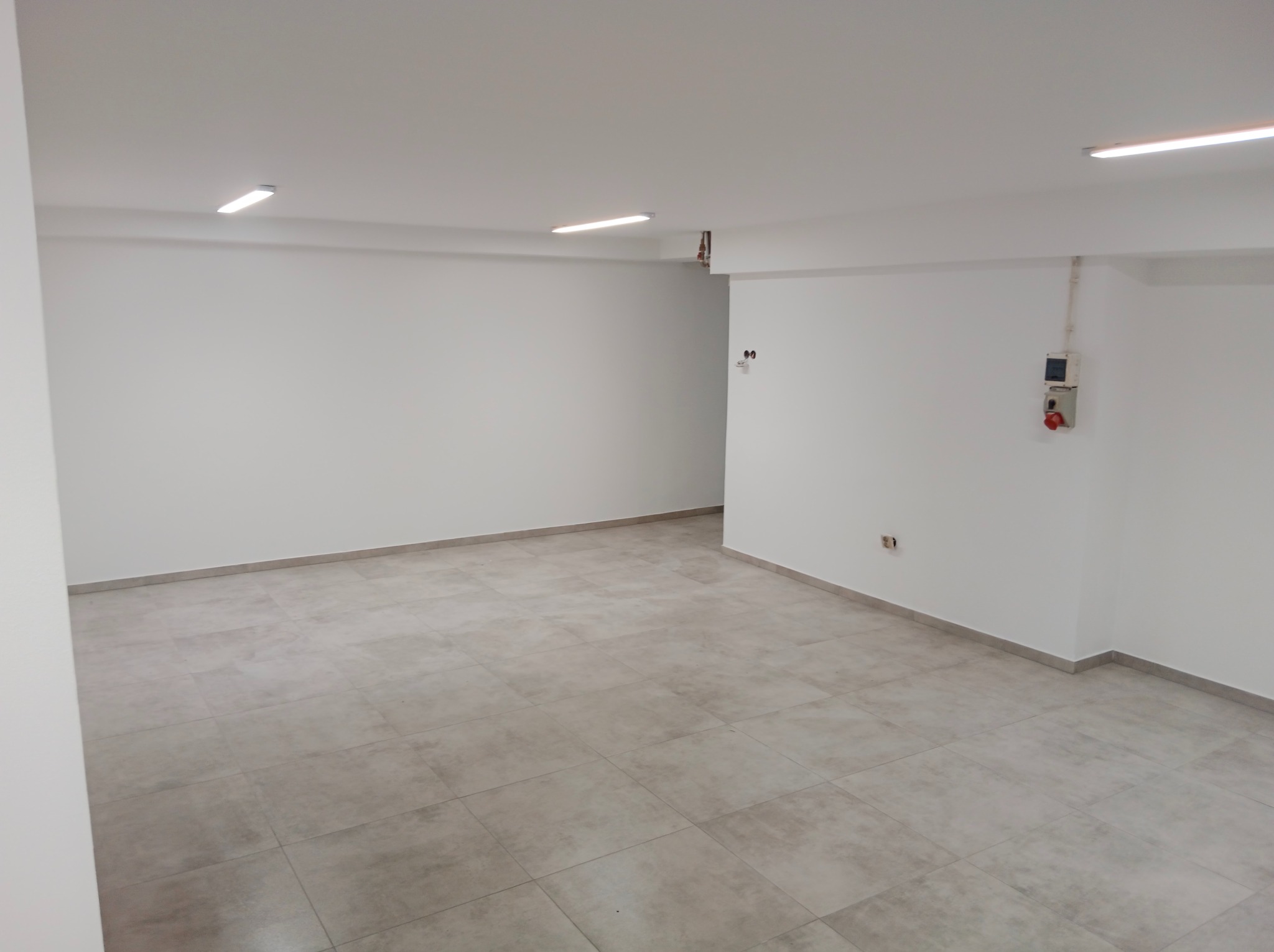 Wykończone wnętrze: jasne płytki podłogowe, świeżo pomalowane białe ściany, widoczne gniazdka elektryczne i oświetlenie sufitowe w pustym pomieszczeniu.