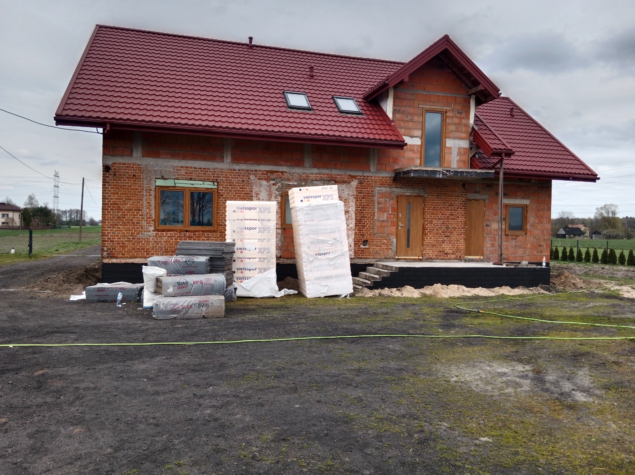 Dom w trakcie budowy z czerwoną dachówką, widoczne materiały izolacyjne XPS. Surowa elewacja z cegły i elementy drewniane okien, okien dachowych i drzwi.