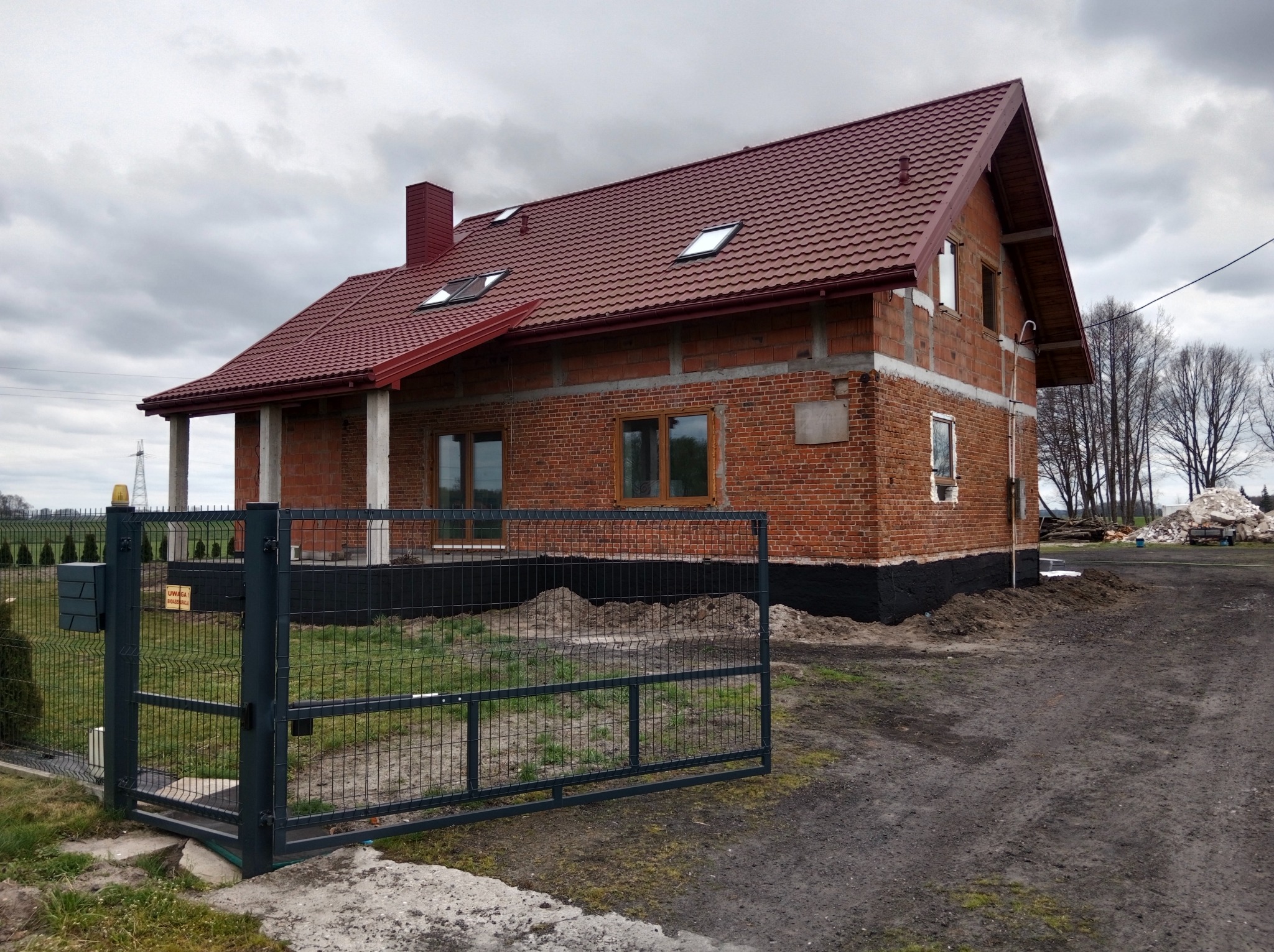 Dom w trakcie budowy z czerwoną dachówką i widoczną, surową cegłą. Otwarta, metalowa brama wjazdowa. Widoczne fundamenty zabezpieczone czarną izolacją.